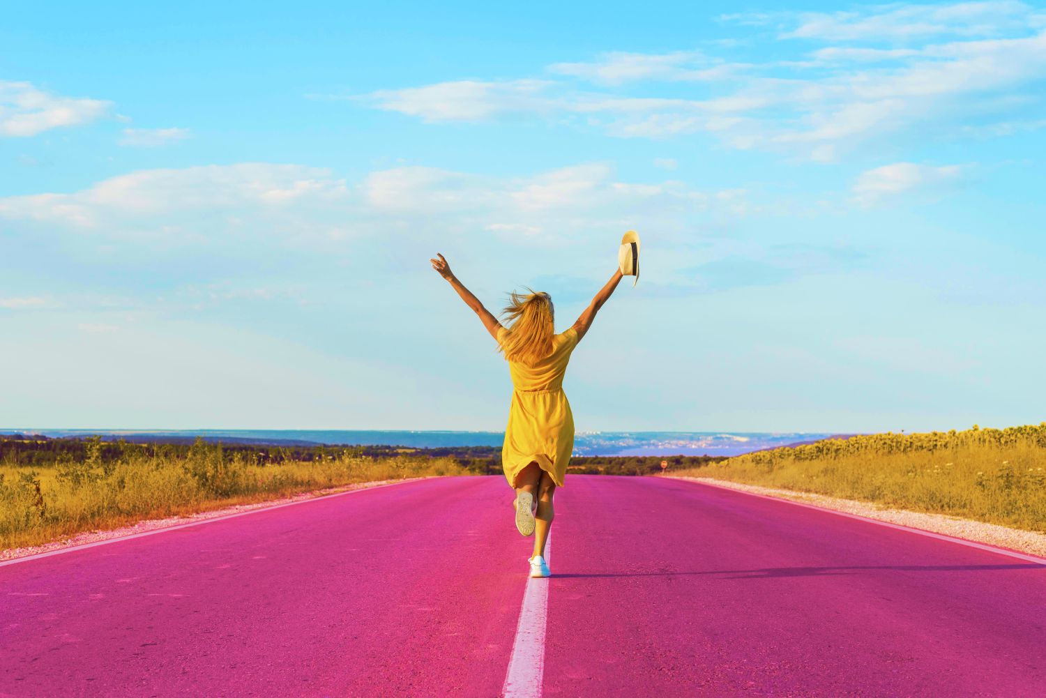 jeune femme qui court sur une route rose, bordée de champs. Elle lève les bras au ciel.cela symbolise la joie de la création de Sens et Pétillance.